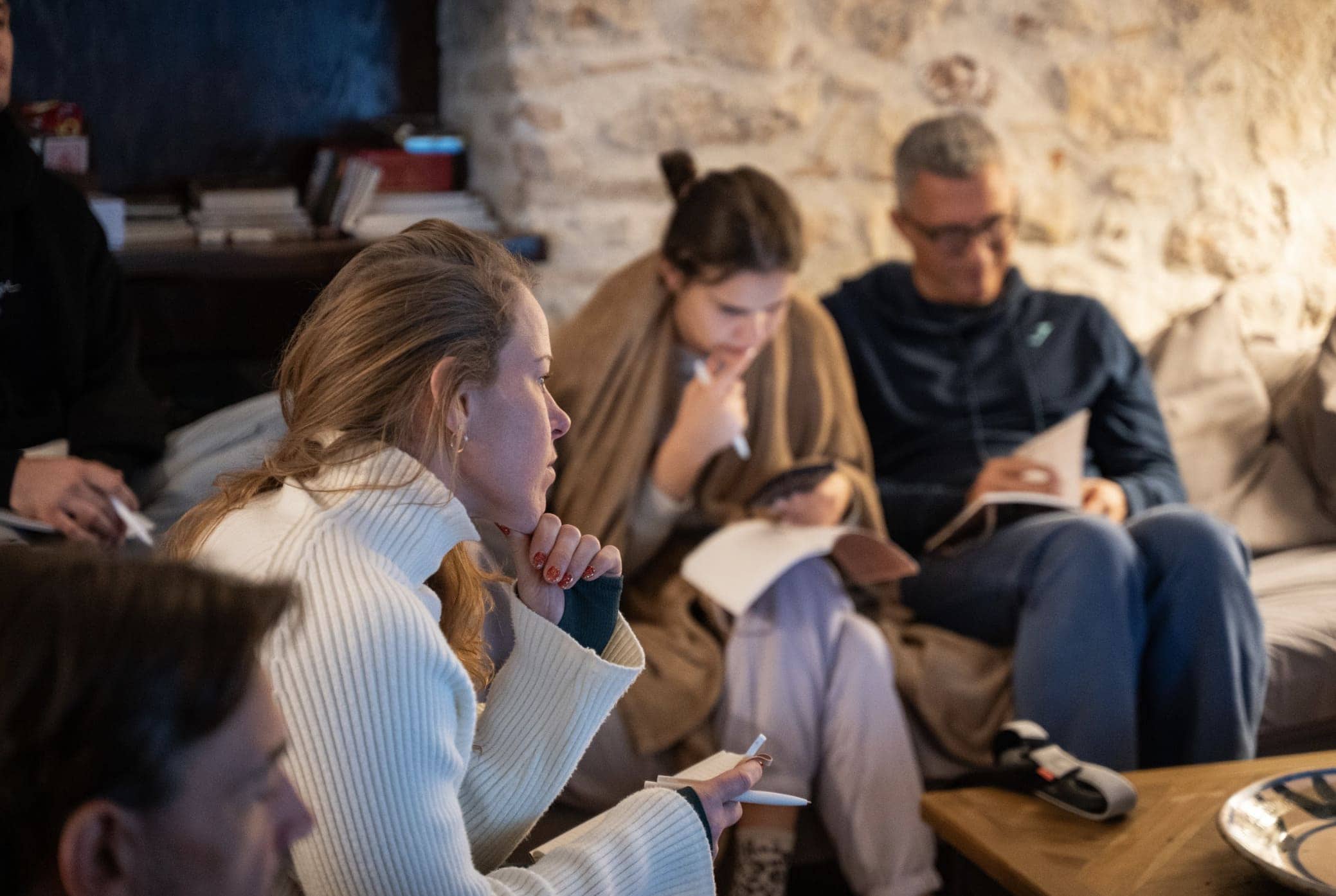 A group of people sit closely together indoors, focused and engaged. Some are holding papers, one checks a phone, and another is deep in thought, suggesting a meeting or discussion in a cozy, stone-walled setting.