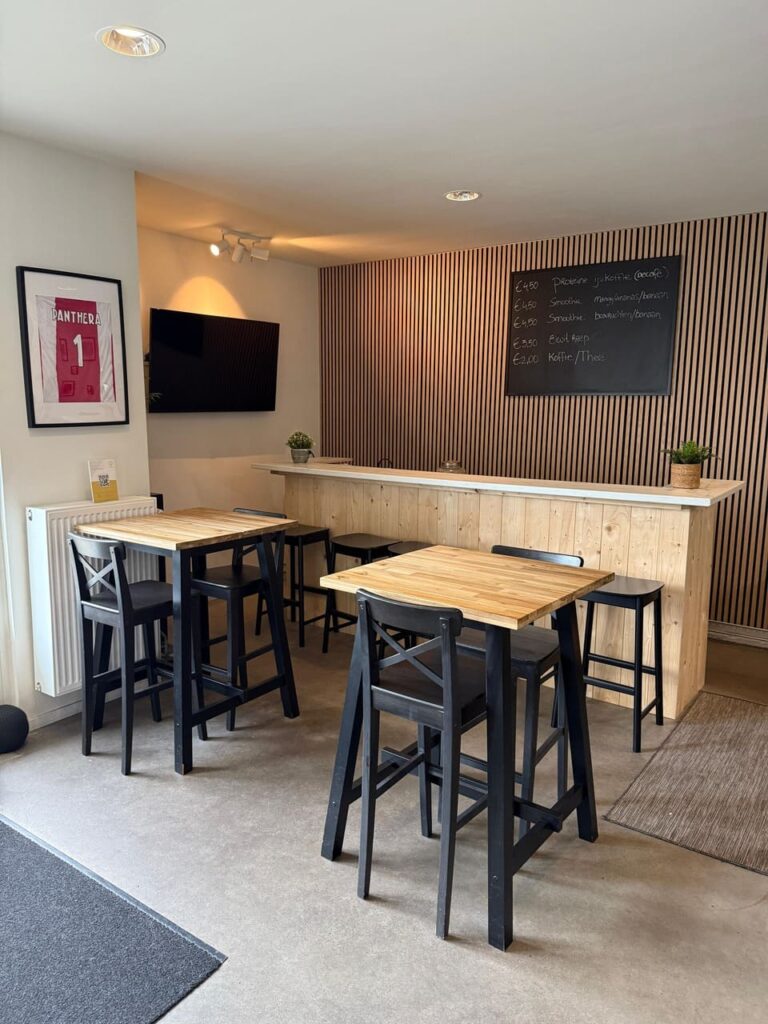 A small café with wooden tables and black chairs, a light wood counter, a menu board on the wall, a TV, framed sports jersey, and potted plants decorating the modern, cozy space.