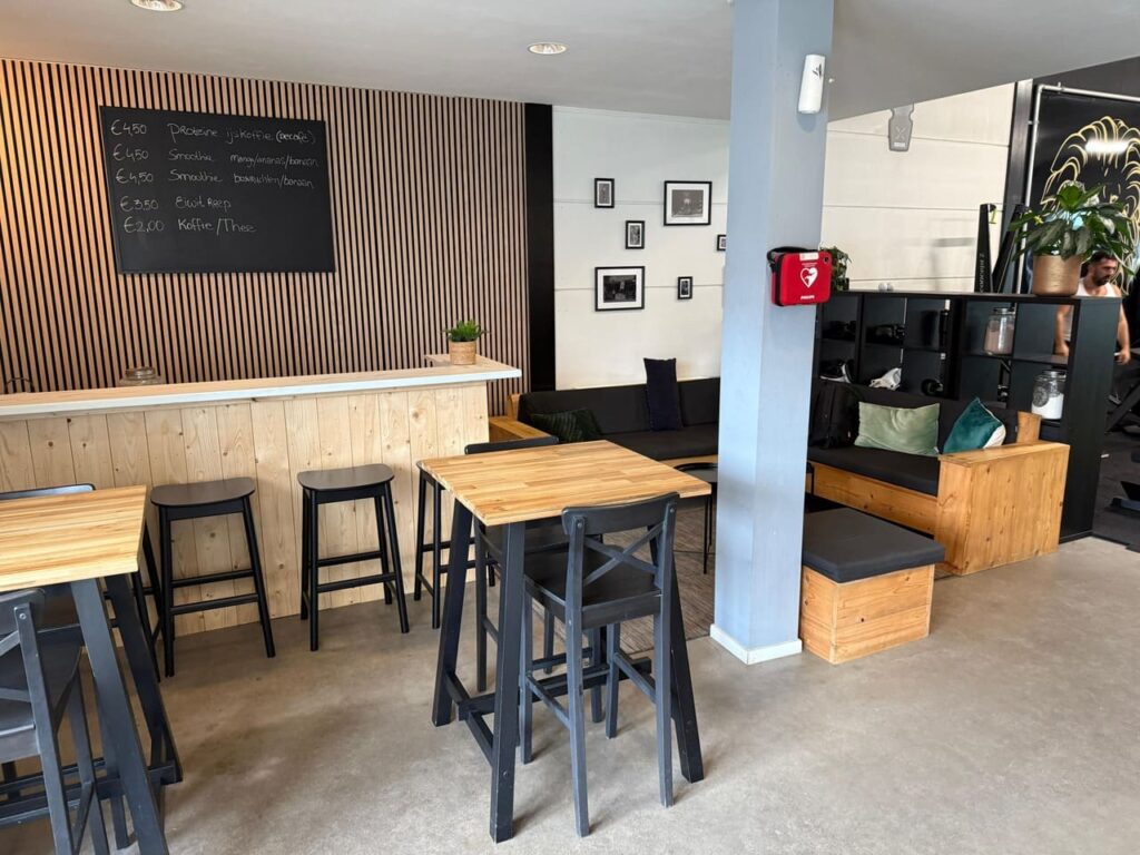 A modern café interior with wooden tables and black stools, a counter with a blackboard menu, black cushioned seating along the wall, and framed photos on a light-colored wall. A fire extinguisher is mounted on a pillar.