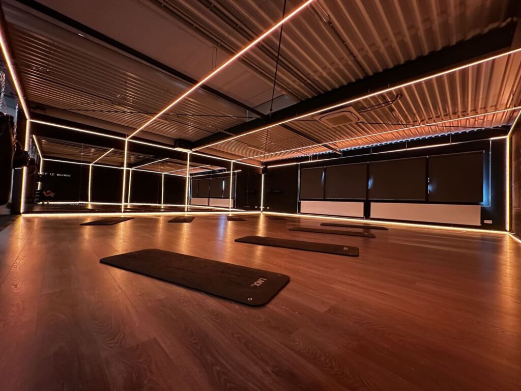 A dimly lit yoga or fitness studio with wooden floors, black exercise mats spaced apart, large mirrors, and warm LED strip lighting outlining the ceiling and walls.