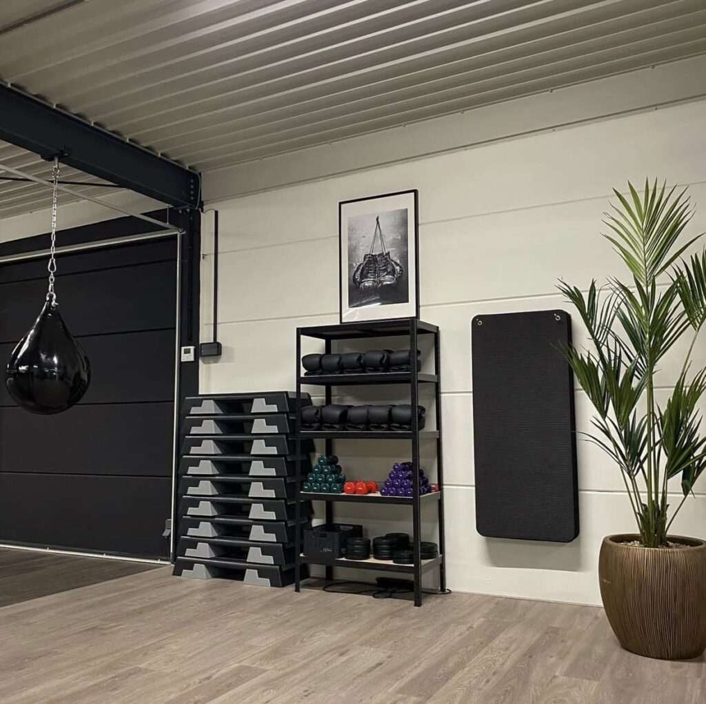 A modern gym room with neatly stacked step platforms, a shelving unit holding yoga mats, dumbbells, and foam rollers, a hanging punching bag, a potted plant, and a framed artwork on a white wall.