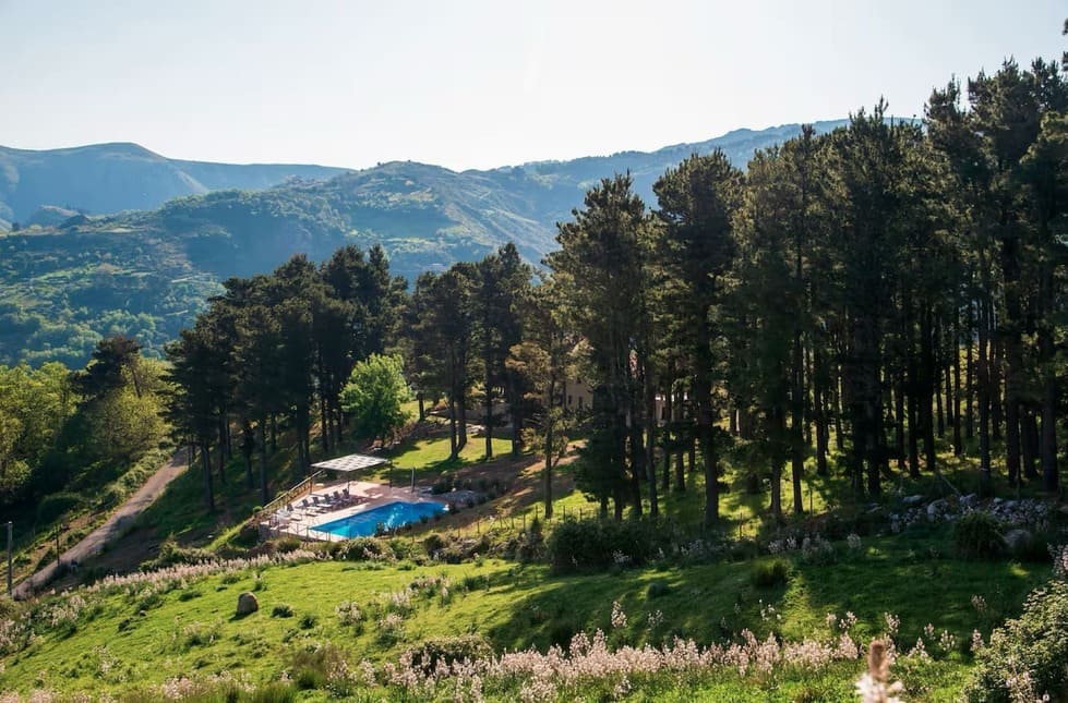 A swimming pool surrounded by trees sits in a green, hilly landscape with mountains in the background under a clear sky.
