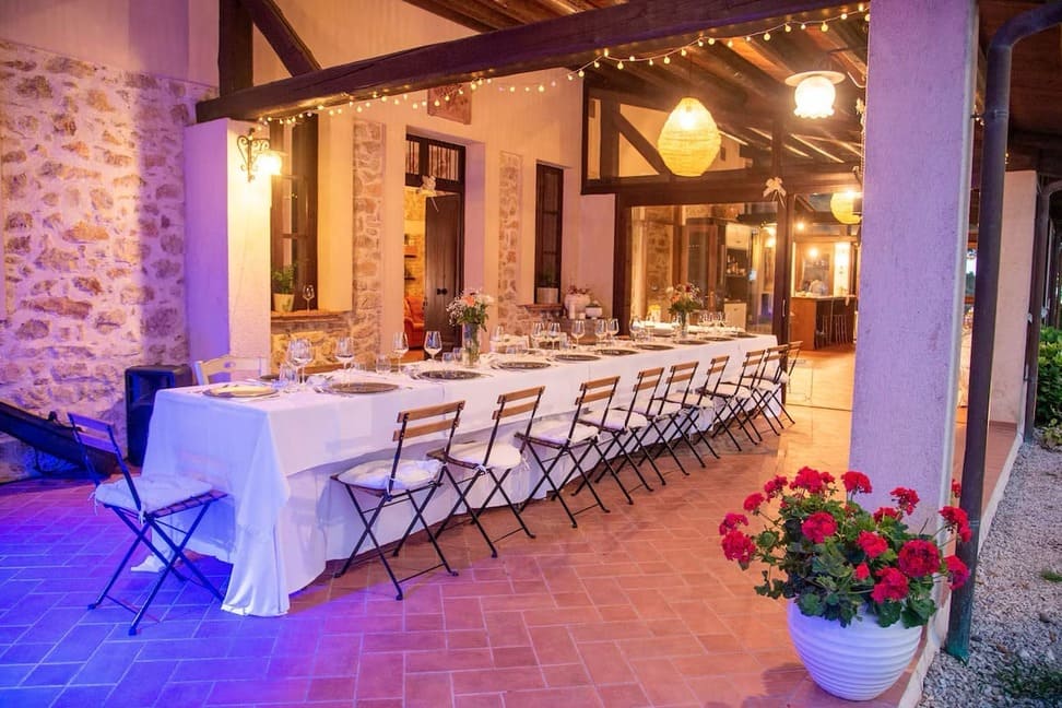 A long table covered with a white tablecloth and set for a formal dinner is arranged on a tiled patio. String lights hang overhead, and potted flowers decorate the space. The setting appears warm and inviting.