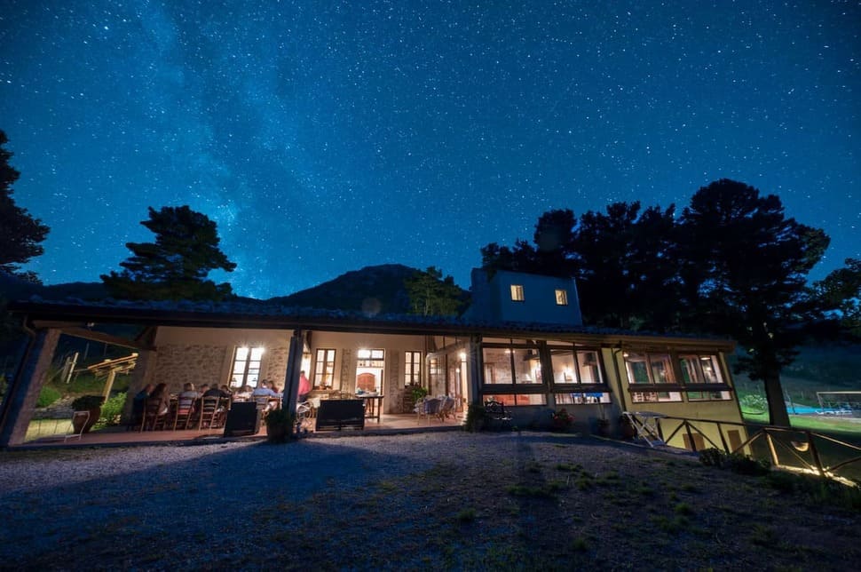 A cozy, well-lit house sits beneath a clear night sky filled with stars, surrounded by trees and hills, with soft indoor lights glowing through large windows.