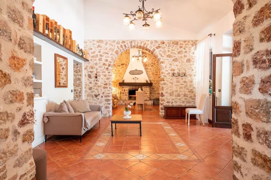 A cozy living room with stone walls, terracotta tile floor, a gray sofa, white shelves, decorative lighting, and an arched doorway leading to a dining area with a fireplace.