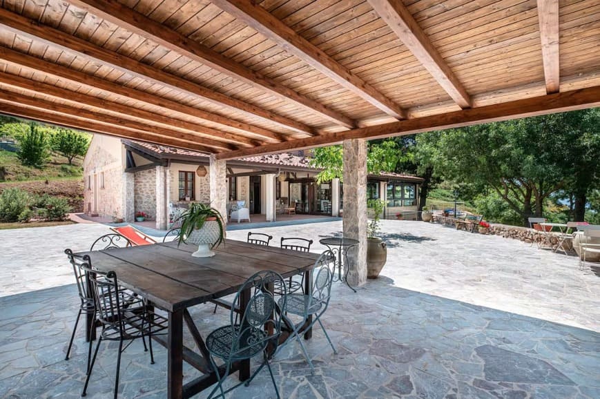 A covered patio with a wooden roof and metal dining table overlooks a spacious stone courtyard, surrounded by a rustic stone house, greenery, and outdoor seating.