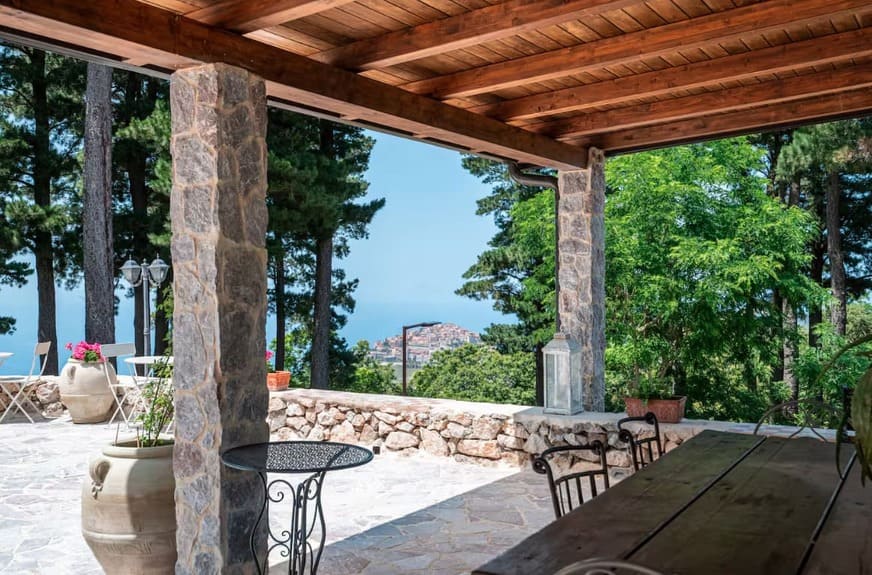A covered stone patio with wooden beams overlooks a scenic view of the sea and distant buildings, surrounded by trees and potted plants, with outdoor tables and chairs on a sunny day.