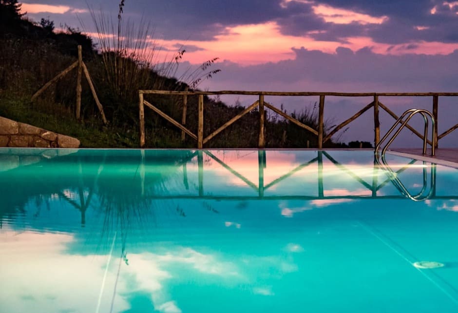 A serene pool reflects a colorful sunset sky with pink and purple clouds. Wooden fencing and tall grass surround the pool, and metal steps are visible on the right side.