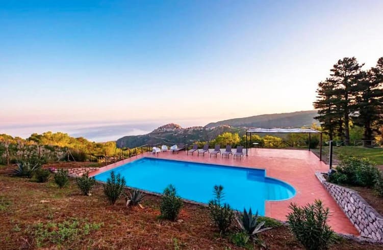 A serene outdoor pool overlooks rolling green hills and a distant town under a clear blue sky at sunset, with lounge chairs lined up along the poolside and lush landscaping surrounding the area.