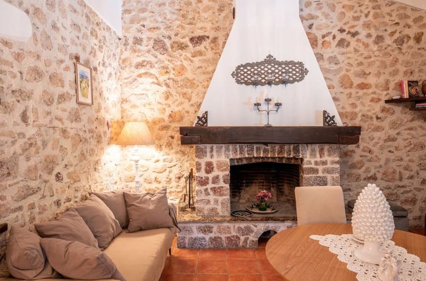 Cozy living room with stone walls, a fireplace with a black mantel, beige sofa with cushions, table with a white decorative centerpiece, lamp, and candlestick, creating a warm, rustic atmosphere.