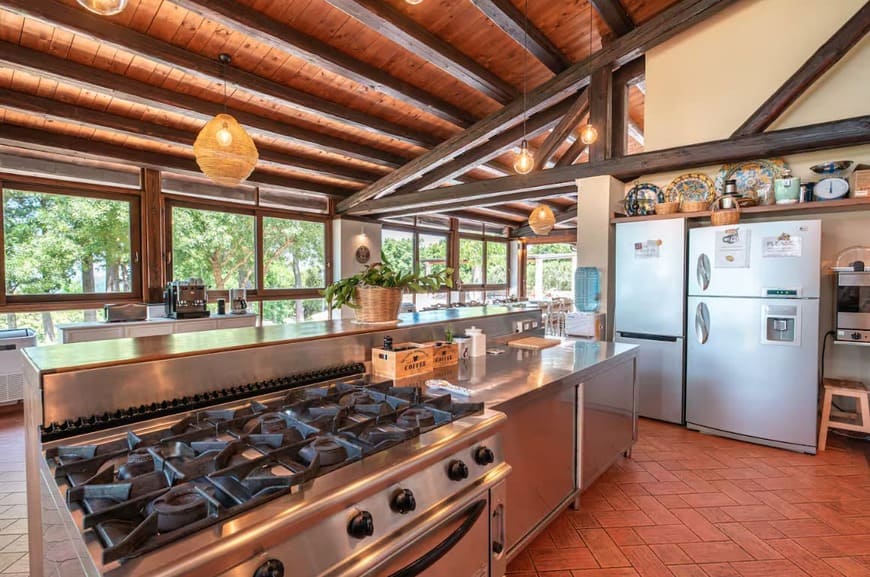 Spacious kitchen with stainless steel countertops, large gas stove, and two refrigerators. Wooden beams and ceiling, terra cotta floor tiles, large windows with views of greenery, and warm lighting.
