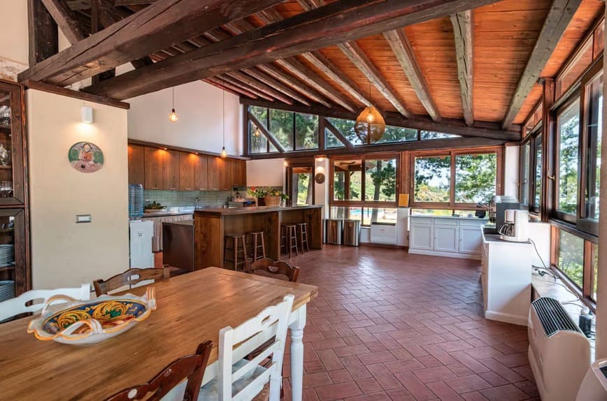 Spacious rustic kitchen and dining area with wooden beams, terracotta tile floor, large windows with forest views, wooden cabinets, and a wooden dining table with white chairs. Natural light fills the room.