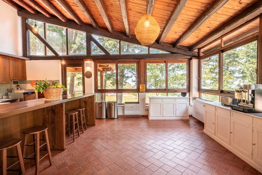 Spacious kitchen with large windows, wooden ceiling beams, terracotta tile floor, wooden bar with stools, white cabinets, and greenery outside. Natural light fills the room, creating a warm, inviting atmosphere.