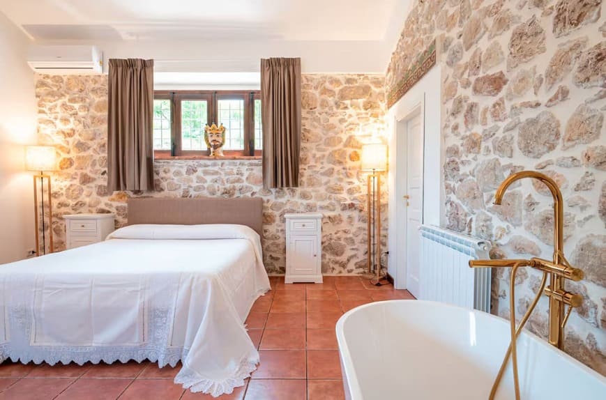 A cozy bedroom with exposed stone walls, terracotta tile floor, a bed with white linens, two bedside tables, a window with brown curtains, and a white freestanding bathtub with a gold faucet in the foreground.