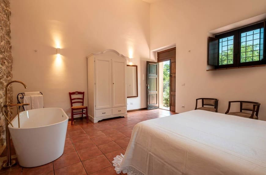 A bright bedroom with terracotta tile flooring, a white bed, a freestanding bathtub, a white wardrobe, two wooden chairs, a red chair, and a window with black shutters letting in sunlight.