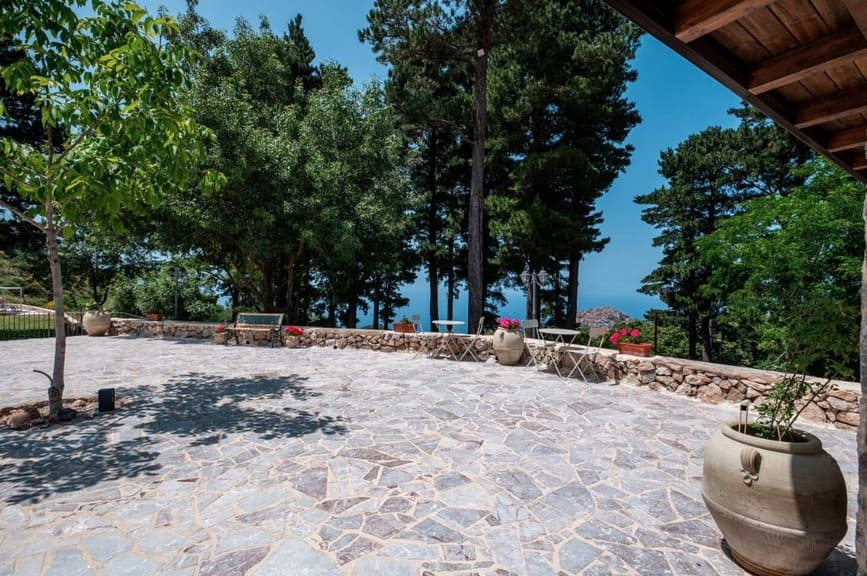 Stone patio with large potted plants, outdoor tables and chairs, surrounded by trees and a low stone wall, with a view of blue sky and distant water beyond the greenery.