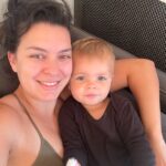 A smiling woman with dark hair and a toddler with blonde hair, both cuddling closely on a gray couch. The toddler is holding a plush toy and they appear relaxed and happy.