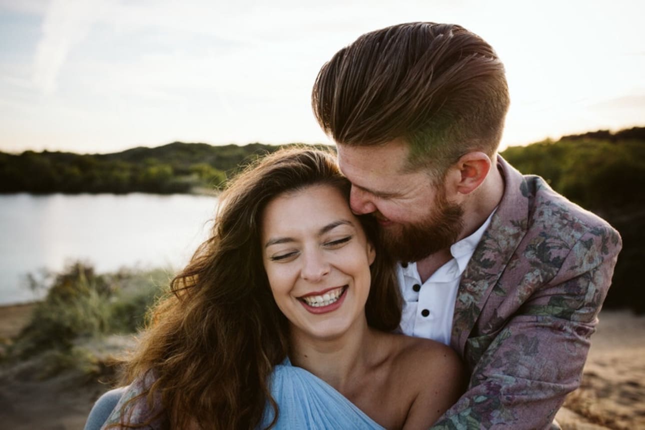 A man hugs a smiling woman from behind near a body of water, both appearing happy and close. The background shows greenery and a bright sky.
