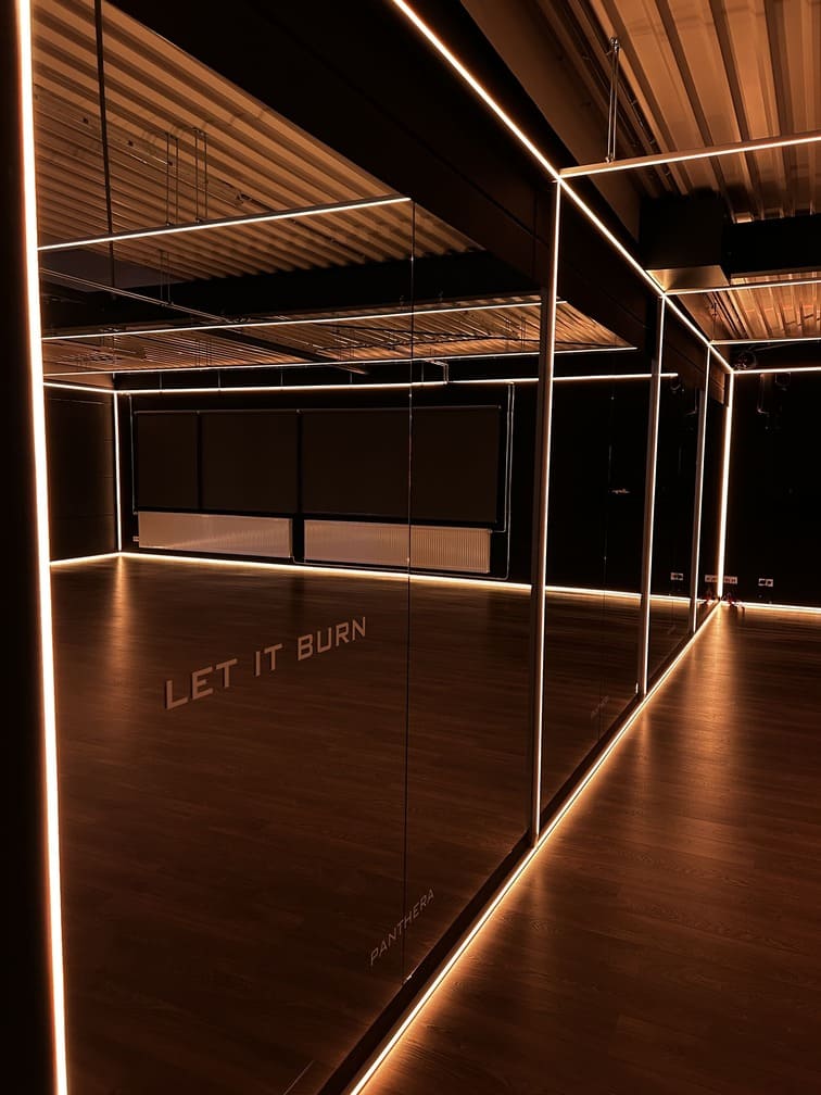 A modern, dimly lit fitness studio with floor-to-ceiling mirrors, black walls, and warm LED strip lights lining the ceiling and edges. The words "LET IT BURN" are reflected on the mirrored wall.