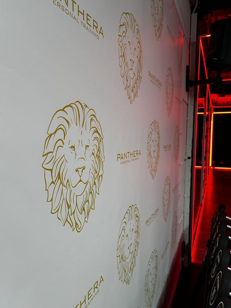 A white wall with repeated gold lion head logos and the text “PANTHERA PERSONAL TRAINING.” A brightly lit gym with red lighting is partially visible on the right side.