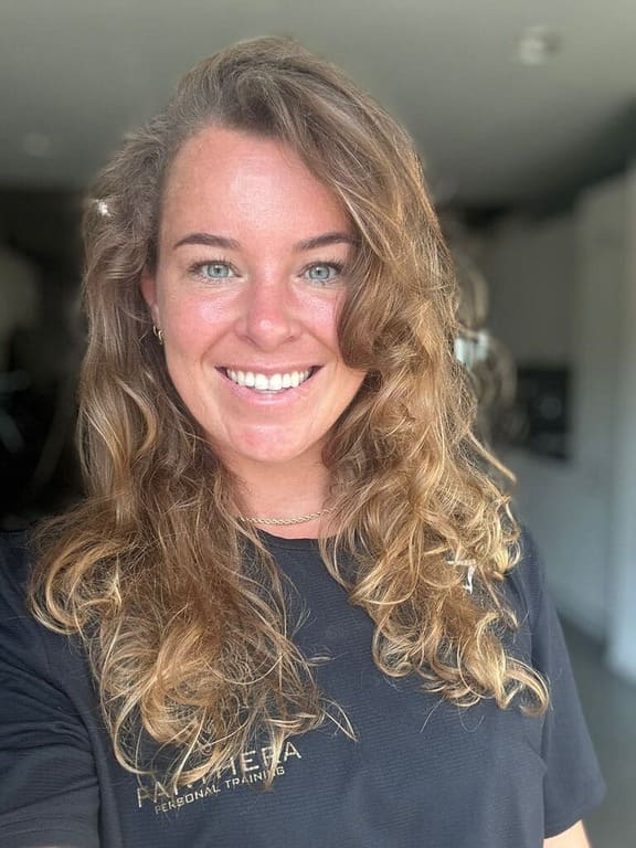 A woman with wavy, light brown hair and blue eyes smiles at the camera. She is wearing a black shirt with gold text and is indoors in a well-lit room with a blurred background.