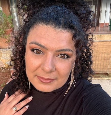 A woman with curly dark hair pulled up wears a black top and long earrings. She has a natural makeup look and is standing outdoors, looking at the camera with a slight smile.