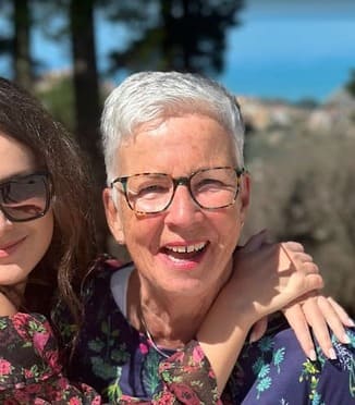 Two women smiling outdoors in sunlight. The older woman with short white hair and glasses is in focus, wearing a floral top, while the younger woman, partly visible, hugs her and wears sunglasses. Trees and blue sky are in the background.