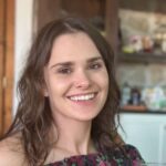 A young woman with wavy brown hair and a nose piercing smiles at the camera. She is wearing an off-the-shoulder floral top. The background shows shelves and a kitchen area out of focus.