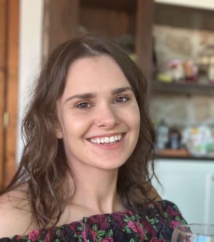 A young woman with wavy brown hair and a nose piercing smiles at the camera. She is wearing an off-the-shoulder floral top. The background shows shelves and a kitchen area out of focus.