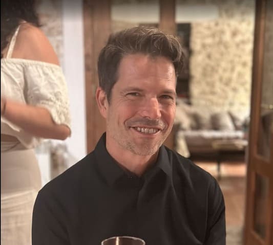 A man with short brown hair and a trimmed beard, wearing a black shirt, smiles while holding a drink indoors. In the background, a woman in an off-shoulder white dress walks by. The room has warm lighting and cozy decor.