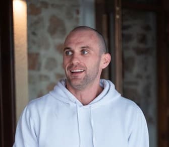A smiling man wearing a white hoodie stands indoors in front of a stone wall and wooden door.