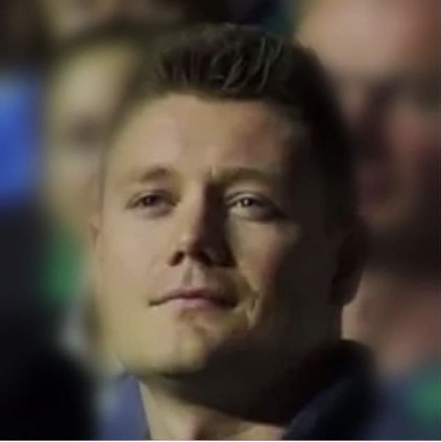 A man with short light brown hair and a slight smile is looking slightly upward. The background is blurred with indistinct people.