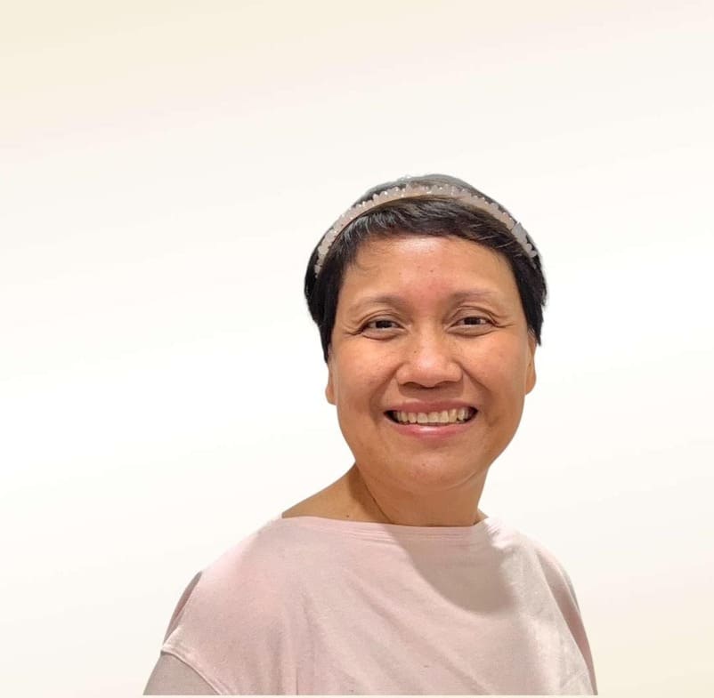 A person with short dark hair smiles warmly, wearing a light pink top and a thin headband, standing against a plain white background.