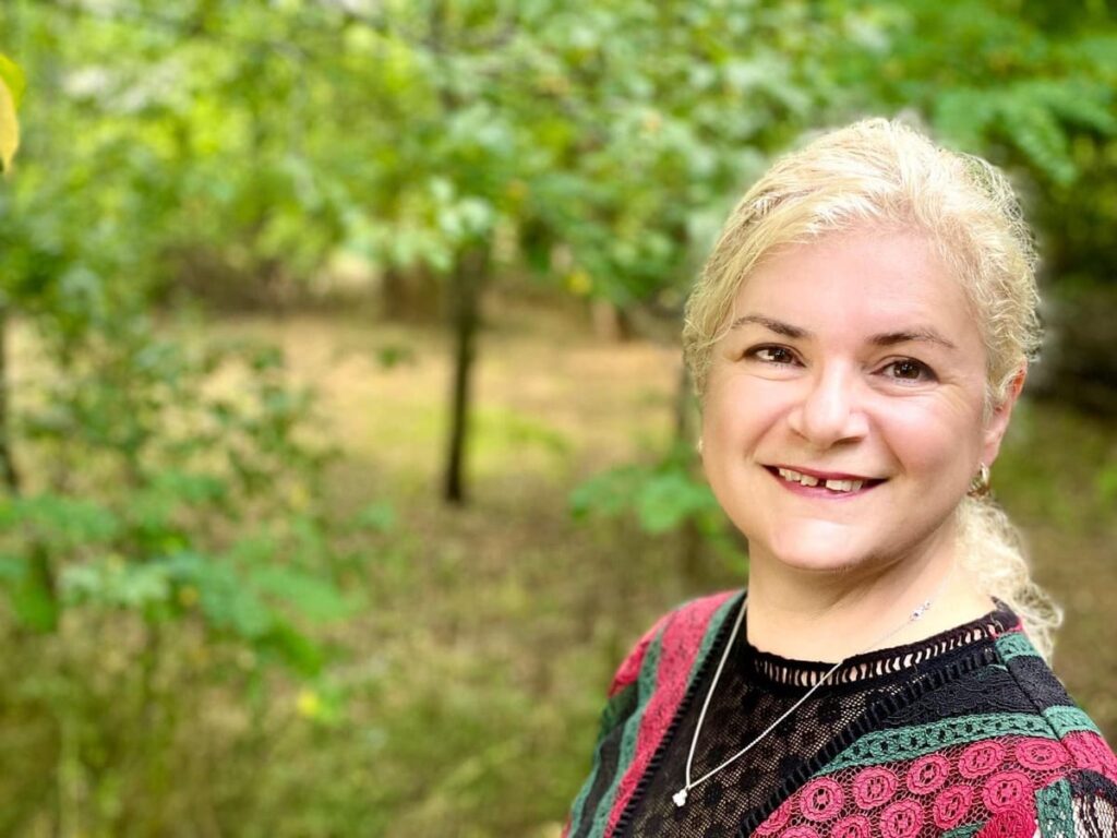 A smiling woman with blonde hair stands outdoors in front of green trees, wearing a black top with red and green patterns and a heart necklace. The background is blurred, highlighting the natural setting.