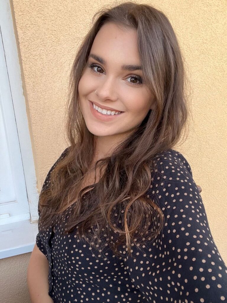 A woman with long wavy brown hair and a nose ring smiles at the camera. She is wearing a black blouse with white polka dots and is standing in front of a beige wall and a white window frame.