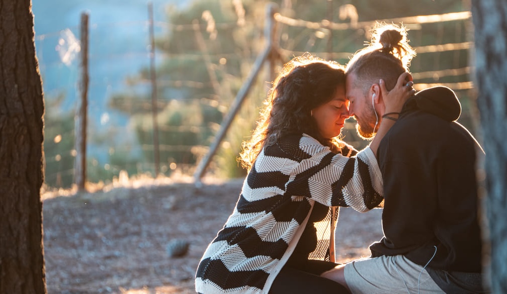 A woman and a man sit close together outdoors at sunset, touching foreheads tenderly. The woman holds the man's head gently, and they appear to share an intimate, comforting moment near a wooden fence.