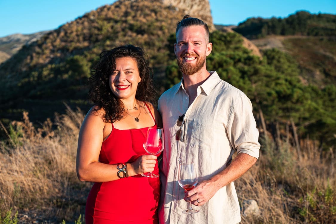 A smiling woman in a red dress and a bearded man in a light shirt stand outdoors holding wine glasses, with hills and greenery in the background on a sunny day.