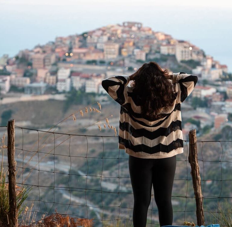 A person with long hair, wearing a striped sweater and black pants, stands facing a hilltop town in the distance, holding their head with both hands. A wire fence and dry grass are visible in the foreground.
