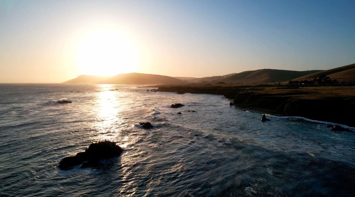 A scenic coastal view at sunset, with sunlight reflecting on the ocean, rocky formations in the water, and rolling hills in the background under a clear sky.