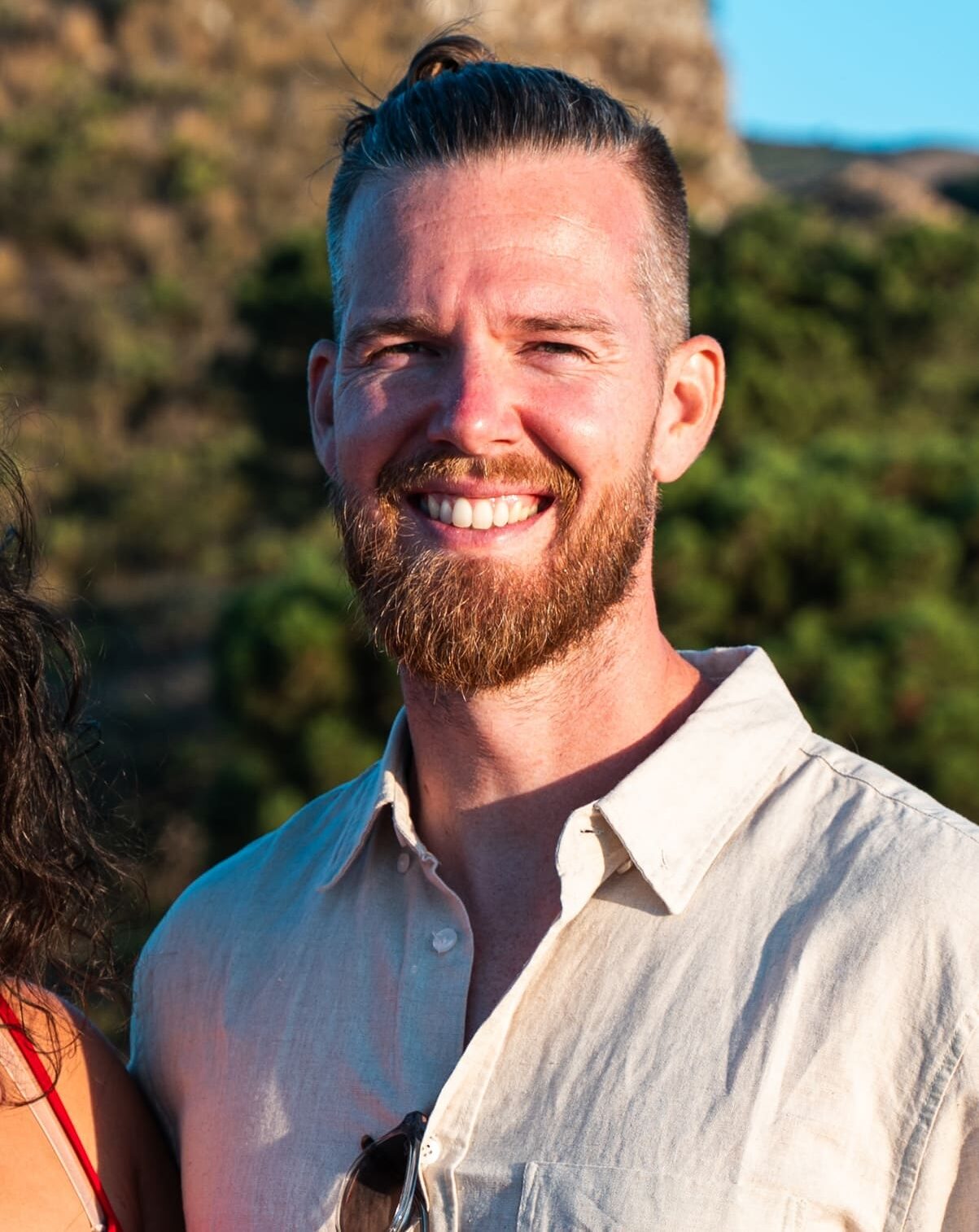 A smiling man wearing a light beige button-up shirt, stands outdoors with greenery and a sunlit hill in the background.