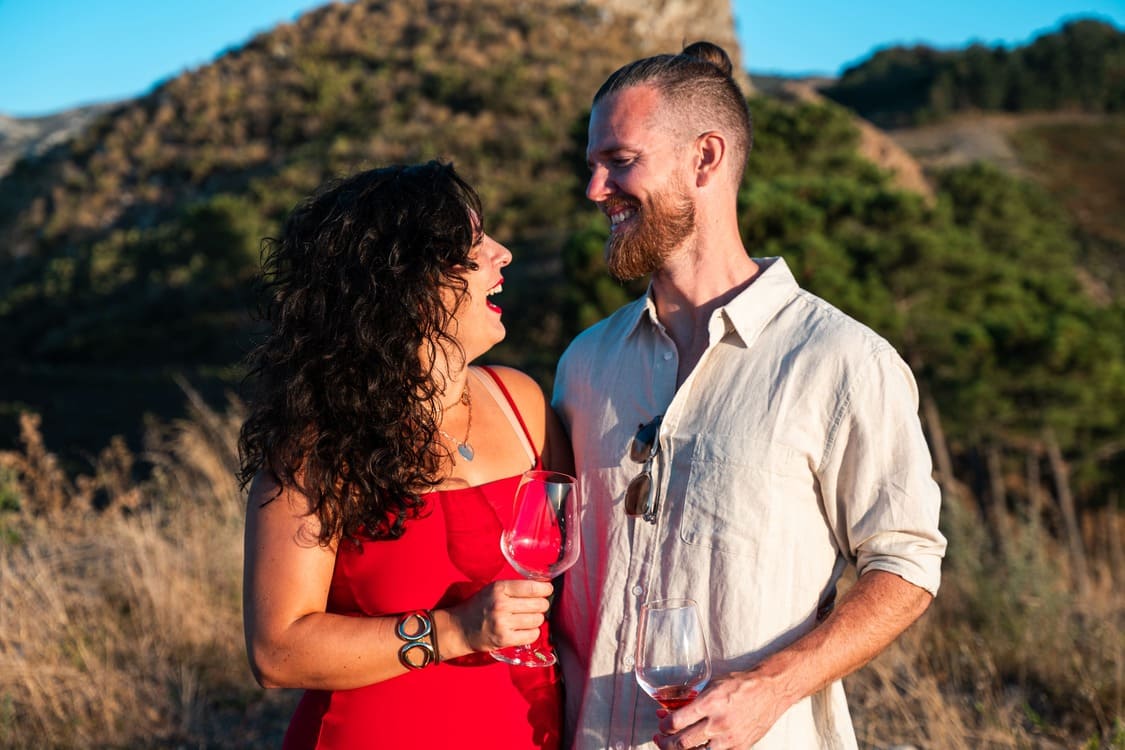 A woman in a red dress and a man in a light shirt stand outdoors, smiling at each other and holding wine glasses, with hills and greenery in the background.