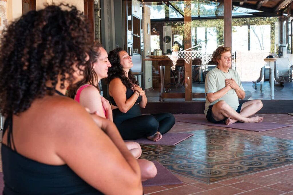 Four people sit cross-legged on yoga mats indoors, eyes closed, and hands on their chests, appearing peaceful and focused, participating in a group meditation or yoga class.