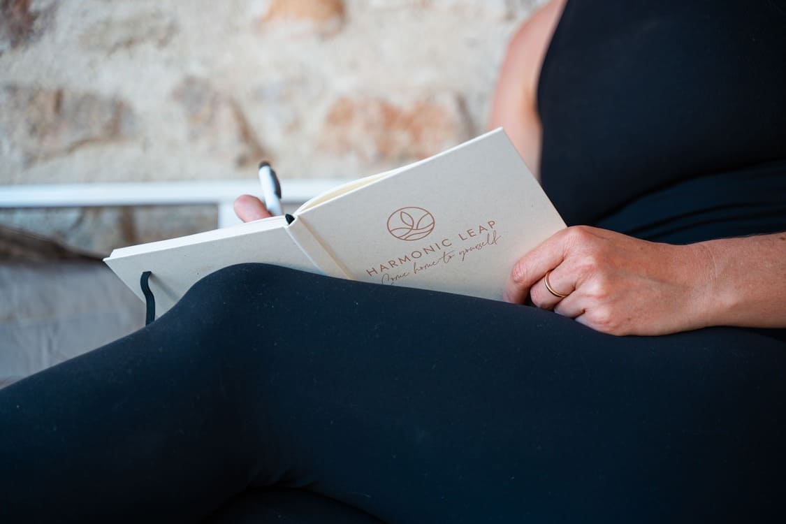 A person wearing black leggings is sitting and holding an open notebook titled "Harmonic Leap" with a pen in hand, appearing to write or journal.