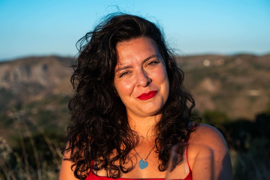 A woman with curly dark hair and red lipstick smiles outdoors, wearing a red top and a heart-shaped necklace, with hills and blue sky in the background.