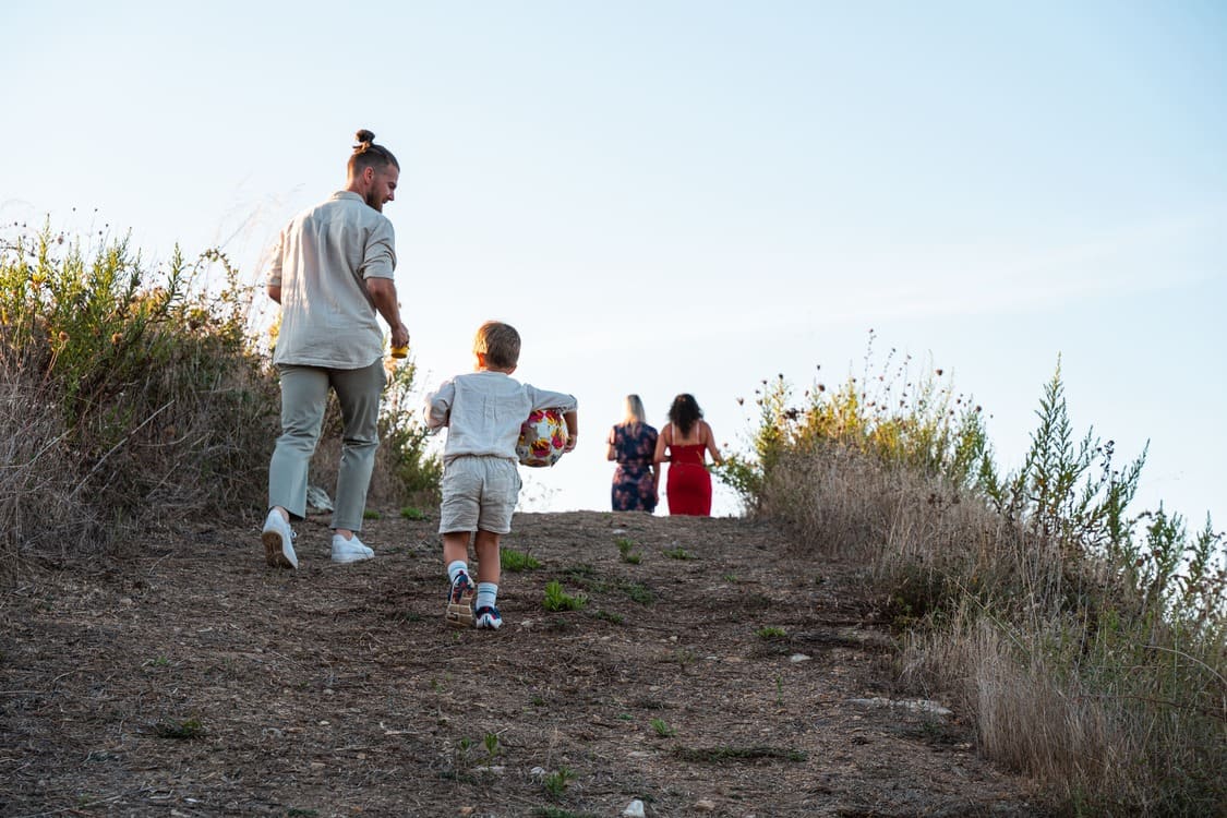 A man and a young boy holding a soccer ball walk up a dirt path, with two women in colorful dresses walking ahead. The path is surrounded by dry grass and shrubs under a clear blue sky.