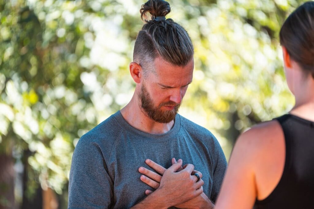 A man with a beard and a top knot holds his hands over his chest, eyes closed, appearing thoughtful or meditative, standing outdoors next to another person.