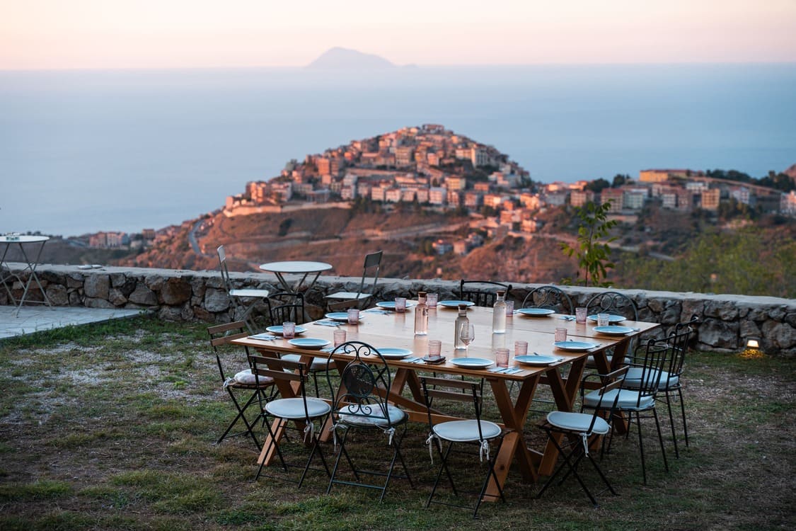 A large outdoor dining table set with plates and glasses overlooks a hillside town and the sea at sunset, with soft lighting and distant mountains in the background.