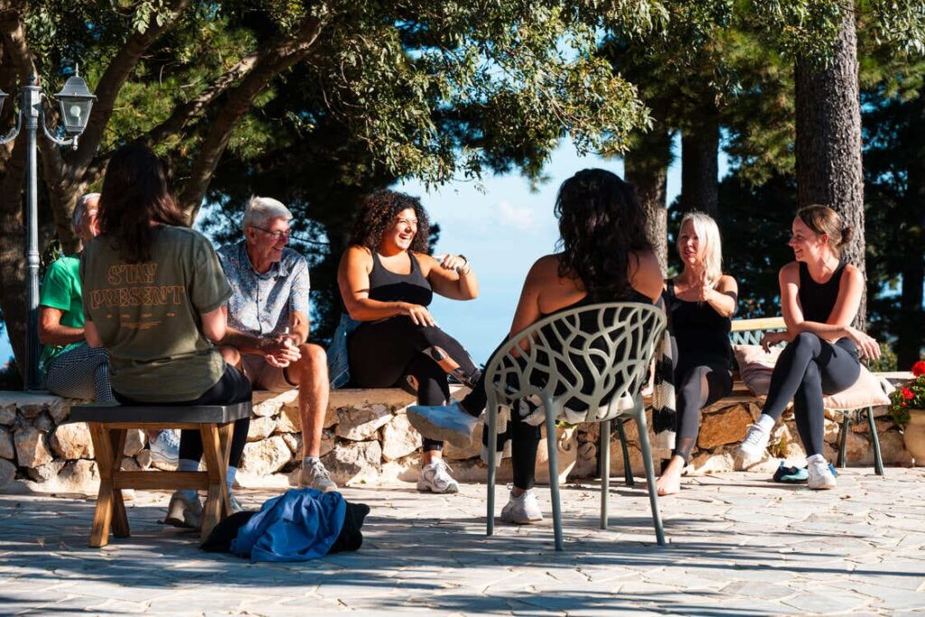 A group of people sit in a circle outdoors on a sunny day, laughing and talking together. They are surrounded by trees and stone walls, creating a relaxed and cheerful atmosphere.