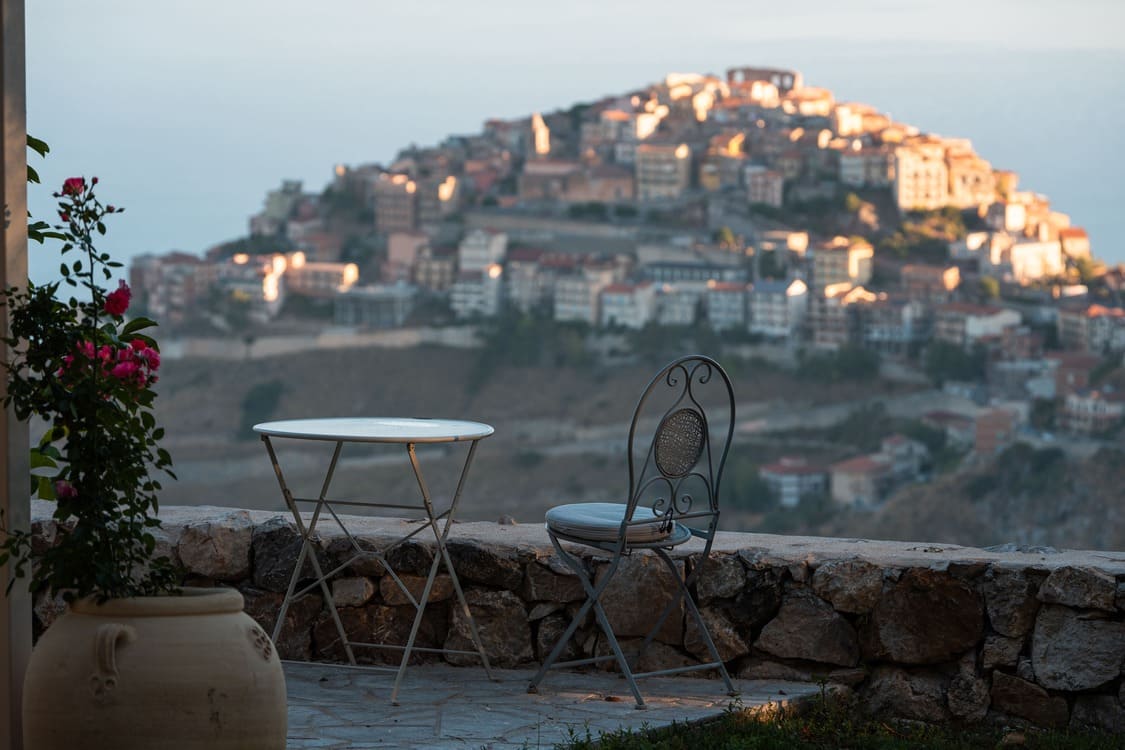 A metal chair and round table sit on a stone patio overlooking a scenic hillside town, with houses clustered on the hilltop and soft sunlight illuminating the view. A potted plant with pink flowers is on the left.