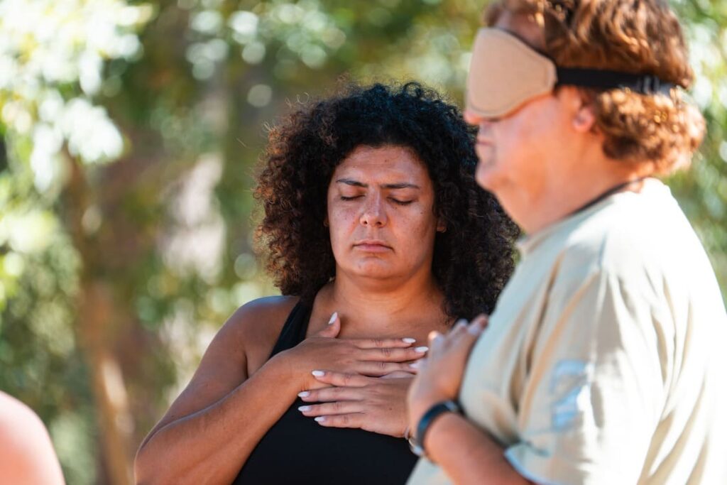 Two people stand outdoors with eyes closed, one with hands on their chest and the other wearing a blindfold. Both appear to be meditating or practicing mindfulness in a natural setting.